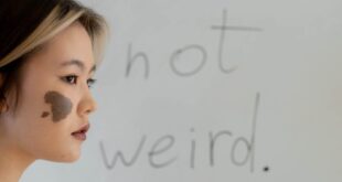 A young woman stands against a whiteboard with the phrase 'I'm not weird' written on it.