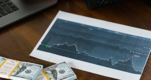 A desk scene with cash bundles, a chart printout, and a person working on a laptop.