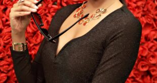 Stylish woman in black dress with sunglasses poses elegantly in front of a vibrant red rose backdrop.