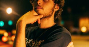 A young man with curly hair poses thoughtfully under city streetlights at night.