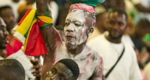 A spirited Ghanaian fan with painted body holding a pot at a crowded soccer stadium, showing team support.