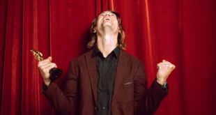 Joyful man in a suit celebrates winning an award on stage with a red curtain backdrop.