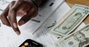 A person calculating expenses using a smartphone and counting cash bills on a wooden table.