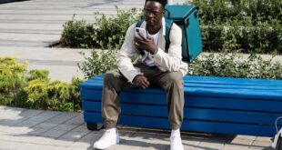 A delivery man sits on a bench outdoors, checking his phone with a thermal bag beside him.