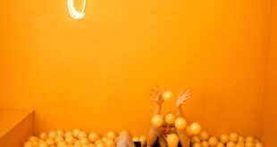Playful scene with a teenager enjoying an orange ball pit beneath a neon 'Juicy' sign indoors.