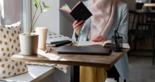 A young woman in a hijab reading a book at a cozy cafe table by the window.