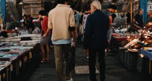 Two men walk through a busy indoor market surrounded by various stalls and goods.