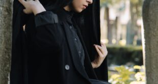 Side view of calm young ethnic female in traditional black clothes with headwear sitting near stone shabby tombstone on cemetery