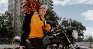 Stylish couple posing with a motorcycle on a dirt road in front of a building.