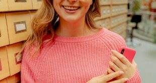 Young woman smiling in a library with a smartphone and eyeglasses, wearing a pink sweater.