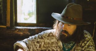 Bearded man in hat sharpening stick at wooden table inside a rustic cabin.