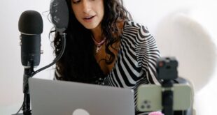 A young woman using a microphone and laptop to record a podcast indoors, showcasing a modern content creation setup.