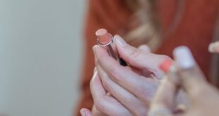 Crop unrecognizable female showing lipstick in hands to friend on blurred background of home