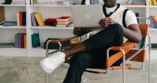 Man sitting in armchair using a laptop in a modern library setting with bookshelves.