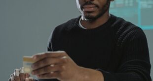 A seated man in glasses and sweater holding a credit card near stacks of cash, suggesting financial themes.