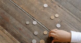 Overhead view of child's hand gathering coins into a glass jar on wooden flooring.