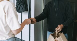 A masked courier hands over shopping bags to a customer in a contactless delivery exchange.