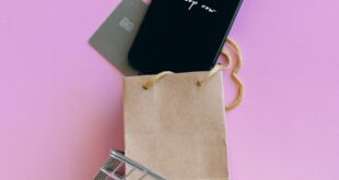 A smartphone, credit card, and bag in a miniature shopping cart on a pink background.