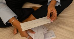 Individual sitting cross-legged examining papers near laptop indoors.