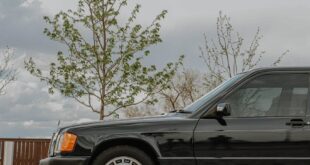 Elegant black luxury car parked outdoors on a paved road with sky and tree background.