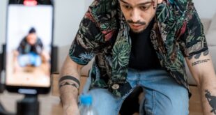 A man filming a bottle flip challenge on a smartphone indoors, capturing a trending social media moment.