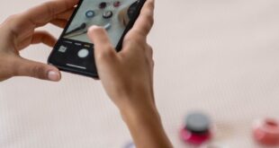A person using a smartphone to capture an image of various makeup products on a bed.