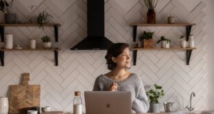Cheerful female in gray sweater sitting at wooden counter with laptop and bottle of milk while browsing internet on netbook during free time at home and looking away