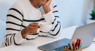 Positive young African American female freelancer in casual clothes talking on smartphone and drinking coffee while working remotely on laptop at home