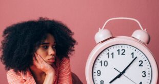 Woman in a pink shirt leans on table with a large alarm clock, conveying impatience.