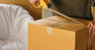 Crop female wearing casual clothes standing while packing carton boxes with scotch tape in modern apartment during preparing for relocation