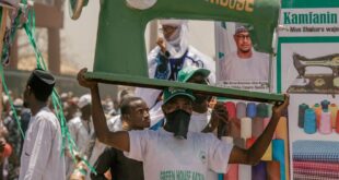 A festive outdoor parade with people carrying a large sewing machine replica and colorful banners.