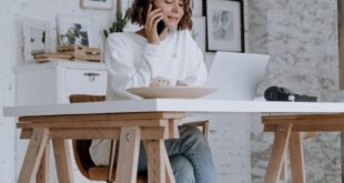 A woman multitasks at home, speaking on the phone while working on her laptop.