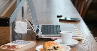 Laptop on a wood table with coffee and pastry in a cozy cafe setting.
