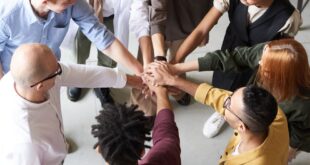 A diverse group of professionals joins hands in a team-building exercise indoors.