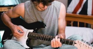 Chilled young adult playing electric guitar on bed, notebook open, Union Jack in background.