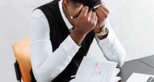 A businessman overwhelmed working indoors with a notebook, laptop, and hourglass.