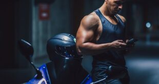 A muscular man checking his phone beside a motorcycle in a dimly lit garage.