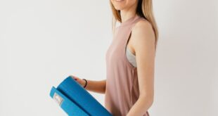 Side view of female athlete in sportswear carrying blue sport mat against white wall and looking at camera over shoulder while preparing for fitness training indoors