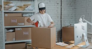 A warehouse worker in uniform packaging boxes for delivery in an organized storage area.
