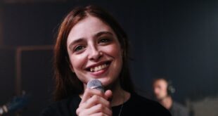 A cheerful female singer performs indoors, holding a microphone with a tattooed arm.