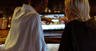 Two women browsing a laptop together at a cozy café bar, seen from behind in a relaxed atmosphere.