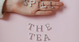 Hand holding 'SPILL' letters with 'THE TEA' on pink backdrop.