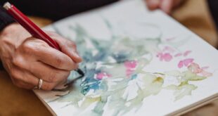 Close-up of hands painting delicate watercolor flowers and foliage in a sketchbook.