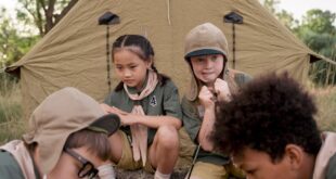 Group of diverse children playing chess outdoors in front of a tent at a scout camp.