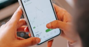 High angle of crop anonymous female broker using app on cellphone with graphic on screen against netbook at home