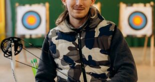 Portrait of a confident male archer holding a compound bow indoors with archery targets in the background.