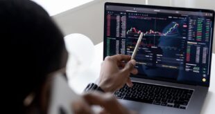 A businessman using a smartphone while analyzing stock market graphs on a laptop indoors.