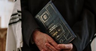 A Jewish man in a synagogue holding a religious text while wearing a tallit.