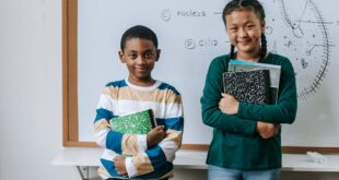 Cheerful schoolchildren holding textbooks in a classroom with a whiteboard drawing.