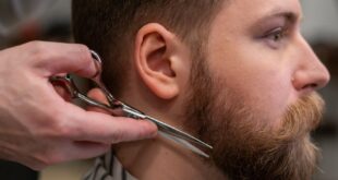 A side view of a barber trimming a bearded man's hair in a barber shop.